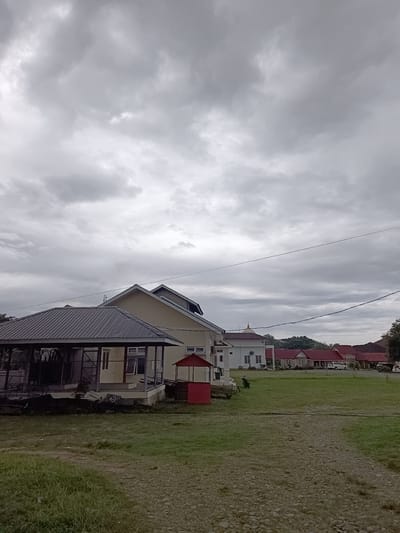 The image depicts a scene in Mesjid Reuntoh, Indonesia, under an overcast sky. In the foreground, a gravel path winds through a grassy field. To the left, a building with a dark metal roof, possibly a pavilion or open-air structure, is visible. Behind it, a light-colored building with a gabled roof is situated. Further in the background, a series of buildings with red roofs line a street, with several vehicles parked along the side. The overall impression is of a somewhat institutional or residential area with a muted color palette due to the cloudy weather. No people are clearly visible in the image.