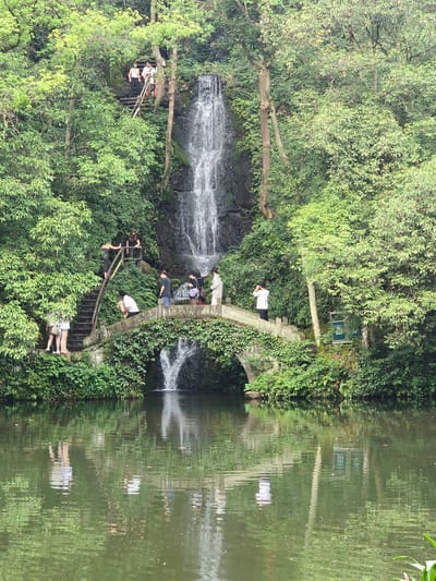 The image depicts a scenic view of a waterfall cascading down a rocky cliff face, surrounded by lush green foliage. In the foreground, a tranquil body of water reflects the surrounding greenery and the stone arch bridge that spans the water. Several individuals are present in the scene, interacting with the environment and each other.
On the bridge, a group of people are gathered. One person, wearing a white shirt and dark pants, appears to be photographing the waterfall with a camera. Another person, in a white shirt, is looking out at the water. To the left of the bridge, a few individuals are standing on a stone staircase leading up the cliff. On the bridge, further to the right, another person is standing and looking towards the waterfall.
Above the bridge, on a wooden platform near the top of the waterfall, a few more people are visible, appearing to observe the waterfall or the surroundings. One person in a white tank top and dark pants is looking down the stairs.
The environment is densely forested with various types of trees and plants, creating a natural and serene atmosphere. The waterfall's mist lightly sprays the surrounding rocks and vegetation. A sign with Chinese characters is visible on the right side of the image, near the water's edge. The overall scene suggests a popular tourist spot within a natural landscape.