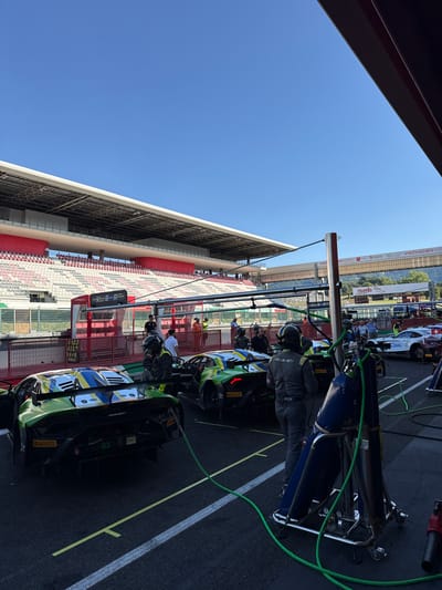 The image captures a scene at the Mugello Circuit, a racetrack located in Scarperia, Italy. In the foreground, two racing cars, one predominantly green with yellow and blue accents and the other white with red accents, are parked in a pit lane. Both cars have sponsor logos, including "IRELLI" visible on the tires and "Lamborghini" on the green car.

Several individuals, dressed in racing attire or team uniforms, are present. Two individuals in the foreground appear to be mechanics or team members, one wearing a grey racing suit and the other a green and black suit with a helmet. One individual in the green and black suit is standing next to a blue gas cylinder on a trolley, which is connected by green hoses to the racing cars. In the middle ground, more team members are visible, some attending to the cars and others observing.

The background features the grandstands of the racetrack, filled with empty red and white seats, indicating the absence of a race in progress or a less crowded event. A clear blue sky dominates the upper portion of the image. Signs with "VISIT TUSCANY.COM" are visible on a banner in the distance, reinforcing the location. The overall scene suggests a preparation or maintenance period for a racing event at the circuit.