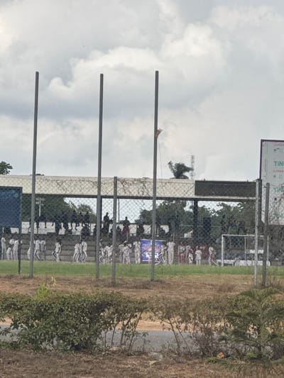 The image displays an outdoor sports complex, likely a stadium or training ground, located in the city of Ife, Nigeria, under an overcast sky.

The foreground consists of dry, brown earth and sparse patches of green shrubbery. Three prominent, evenly spaced, vertical gray metal poles rise from behind this vegetation, extending upwards into the frame. A chain-link fence runs horizontally across the mid-ground, acting as a barrier.

Behind the fence, a grassy field is populated by numerous individuals, predominantly appearing as children or young adults. Many are dressed in matching white or light-colored uniforms, with some wearing red sashes or accents. They are dispersed across the field, with several groups appearing to be in formations or moving in lines. A blue banner with white text is visible on the field to the left of the center. A white metal goalpost structure is partially visible on the far right of the field.

In the background, a concrete grandstand or seating structure is visible, occupied by additional individuals seated or standing. Beyond the grandstand, trees and green foliage are present. To the far right, a large billboard or sign displays the text "TIN" in white letters on a light background. A metal lattice communication tower is discernible in the distant background, rising above the grandstand structure on the right side of the frame.