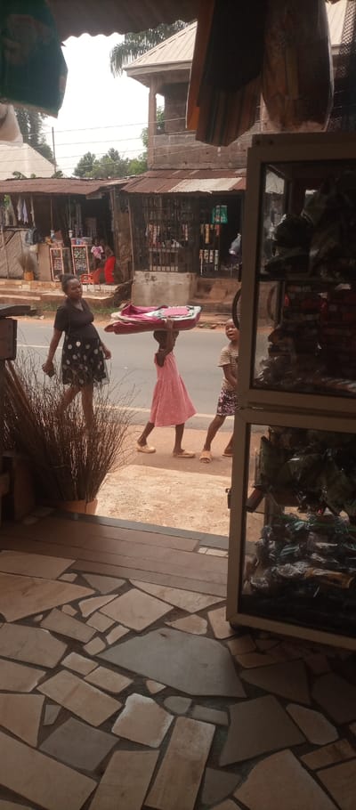 The image captures a street scene in Umuoji, Nigeria, viewed from the interior threshold of a commercial building. The foreground displays a floor constructed from irregularly shaped light-colored flagstones with dark grout. To the left, a tall brown planter holds dried, thin brown grasses. To the right, a display cabinet with a light-colored frame and glass shelves contains numerous individually wrapped dark items.
In the midground, a paved asphalt road is bordered by a lighter, unpaved dirt shoulder. Three individuals are walking on this shoulder from left to right. An adult female, dressed in a black V-neck top and a black skirt with white polka dots, carries a dark smartphone in her right hand. Directly behind her, a young girl in a pink polka-dot dress balances a folded, reddish-brown fabric object on her head. To the girl's right, partially obscured by the display cabinet, another child wearing a striped top and patterned skirt appears to look towards the left. All three individuals are wearing light-colored sandals or flip-flops.
Across the asphalt road, a series of single-story structures with rusted corrugated metal roofs are visible, functioning as shops or market stalls. These structures display various goods, including white shirts hanging for sale and packaged items on shelves behind metal grates. A person in a red garment is discernible further within one of these distant establishments. The background features a more substantial two-story building with a light-colored roof and exposed blockwork, partially obscured by trees and palm fronds. The sky overhead is bright and hazy.