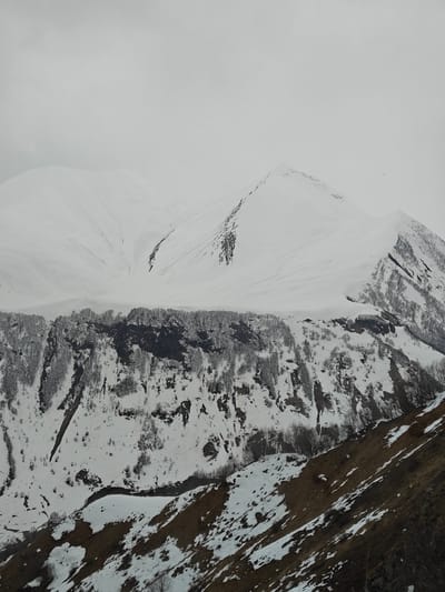The image shows a mountainous landscape covered in snow, likely located in the Dariali region of Georgia. The foreground consists of a brown, grassy slope with patches of snow. A river runs through this area. Above this, there's a steep, rocky cliff covered with trees, some with snow on their branches. The background is dominated by a large, snow-covered mountain range under a cloudy sky. The snow appears deep and continuous on the mountain peaks.