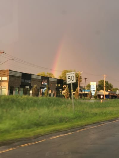 The image captures a scene on a road in Saint-Jean-sur-Richelieu, Canada, under a twilight sky marked by a faint rainbow. On the left, a low-rise commercial building with a brick facade and dark-colored accents is visible. Several windows and doorways are present along its front. To the right of the building, a grassy verge separates it from the road, which features yellow lane markings. Further to the right, utility poles with overhead wires are present. A speed limit sign indicating "50" is prominently displayed. Beyond the speed limit sign, more low-rise buildings and trees are visible, with a glowing sign that appears to read "QUAZI." The overall atmosphere is subdued, with the natural light suggesting either sunrise or sunset.