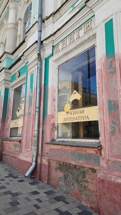 The image shows the facade of a building in Smolensk, Russia, with a sign in the window reading "Учебная Литература" (Educational Literature). The building's facade is characterized by peeling paint and a weathered appearance. The windows are framed with decorative trim. A metal drainage pipe runs horizontally along the facade. The street is paved with gray brick.