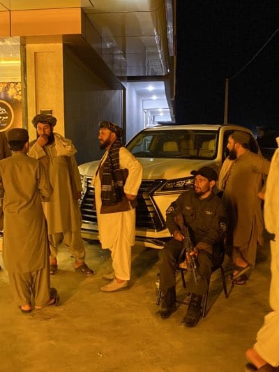 Outside a modern building at night in قندهار, Afghanistan, a group of men stands near a white Lexus SUV. Several of the men wear traditional clothing and head coverings. One man, dressed in a dark uniform and seated on a chair, holds a rifle.
