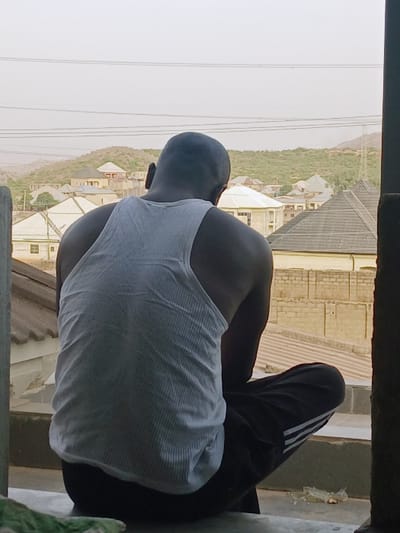 A person, observed from behind, is seated or squatting on a light-colored concrete ledge, looking outwards. The individual wears a white ribbed tank top and dark pants, with white stripes visible on the right pant leg. The scene is located in Kwekwe, Zimbabwe. The background consists of a dense residential area featuring numerous single and multi-story houses with various roof materials, including dark tiles and light-colored concrete. Several structures exhibit exposed concrete block walls. In the distant background, hills with sparse vegetation are visible under a hazy sky. Multiple horizontal power lines traverse the upper portion of the frame. A fragment of green fabric is visible on the ledge in the lower-left, and a small piece of debris lies on the ledge to the lower-right.