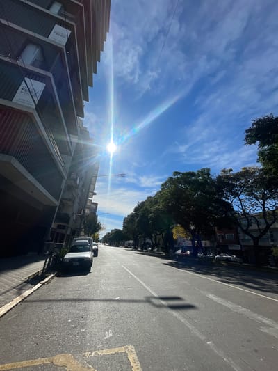 The image shows a street scene in La Lucila, Argentina. The sun is shining brightly in a blue sky with streaky clouds. On the left is a multi-story building with a stepped facade. A sign is visible on the building. On the right is a street with parked cars along the side. Trees line the street, casting shadows. In the distance, more buildings and cars are visible.