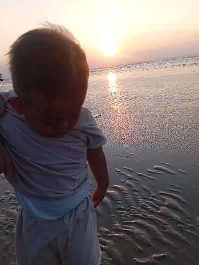 The image captures a baby or young toddler on a beach at sunset, specified to be in the city of Tritunggal, Indonesia. The child, with light brown hair and dressed in a light-colored t-shirt and shorts, is seen from a high angle, partially in the foreground, looking downwards towards the sand. An unseen individual's hand is visible supporting the child's left arm. The background features a vibrant sunset, with the sun prominent and low over the horizon, casting a bright orange and yellow light across the sky. Sunlight reflects intensely on the calm water surface and the wet, dark sand, which displays clear ripple patterns. A dark, indistinct structure is faintly visible over the water in the far left background.