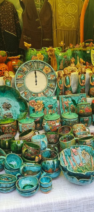 This is a medium-shot, eye-level, vertical-orientation photograph taken at an outdoor market in Старые Быги, Russia. The image displays a variety of handcrafted ceramic items laid out on a white textured tablecloth. The items include numerous bowls, cups, teapots, and decorative plates, predominantly in shades of teal and green with intricate floral and geometric patterns. A large, teal-colored Roman numeral clock with detailed imagery on its face is prominently featured in the center of the frame. Behind the ceramics, several pieces of clothing are visible, including a brown vest and a patterned fabric. The overall setting suggests a craft fair or market stall.