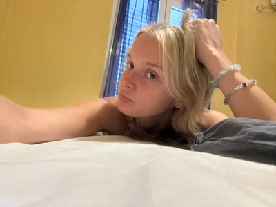 The media file shows a young woman with blonde hair and freckles, lying on a bed. She is wearing a gray t-shirt and several bracelets on her wrist. The room has yellow walls and a window with blue blinds. The lighting suggests it is daytime. The woman appears relaxed and is looking at the camera. The image is shot from a low angle, making it seem as if the viewer is looking up at her. The overall mood is casual and intimate. There is no visible text in the media file. The setting appears to be a bedroom in Arkadii Municipal Unit, Greece.