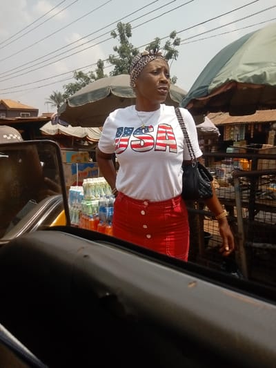The image displays an adult female standing in an outdoor market environment in Umudioka, Nigeria, captured from the perspective of a seated observer inside a vehicle. The subject wears a white t-shirt with a stylized "USA" flag design, a red button-front skirt, a patterned headscarf, a necklace, and carries a black shoulder bag with a chain strap. A gold-colored bracelet is visible on her left wrist. Her head is turned to the right, and her mouth is slightly open. Directly behind her, a market stall features multiple stacks of bottled beverages, including "OAT MILK," shaded by a large, dark green market umbrella. In the background, additional market stalls with corrugated metal roofs, a building with a reddish-brown roof, trees, and overhead power lines are visible against a pale sky. The foreground includes a dark, curved interior surface of the vehicle.