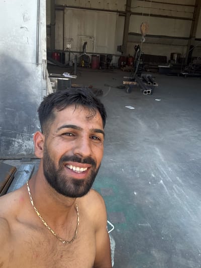 A shirtless man with dark hair and a beard smiles at the camera. He is wearing a gold chain. The man is standing in what appears to be a warehouse or industrial building. In the background, there are various metal objects, including pipes and beams, as well as a crane. The floor is concrete and there are some red barrels visible. The location is the city of Tsaratsovo, Bulgaria.