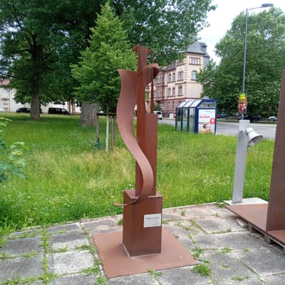 The image shows a rusty metal sculpture in the foreground, standing on a square metal base which is placed on a paved area. The sculpture is abstract, with a tall, rectangular central element and a curved, ribbon-like piece that wraps around it. A small white plaque is attached to the front of the rectangular element, reading "Gedanken an Elisabeth" (Thoughts of Elisabeth), "2004" and "Eduard Schönleber."

Behind the sculpture, there is a large, grassy area with scattered white wildflowers. In the middle ground, a street is visible with cars and a bus stop shelter made of glass and metal. A lamppost with a speaker is positioned to the right of the sculpture. Beyond the street, there are buildings, one of which is a multi-story building with a mansard roof. Tall trees with lush green foliage are scattered throughout the scene. A poster for "Circus Fratelli" is attached to the lamppost.

The scene appears to be outdoors in a public space, likely a park or a plaza in the city of Eisenach, Germany, as indicated by the poster for Circus Fratelli in Eisenach. The overall atmosphere is calm and somewhat urban, with a mix of nature and man-made structures. There are no people clearly visible in the image, except for a possible faint reflection in the bus stop.