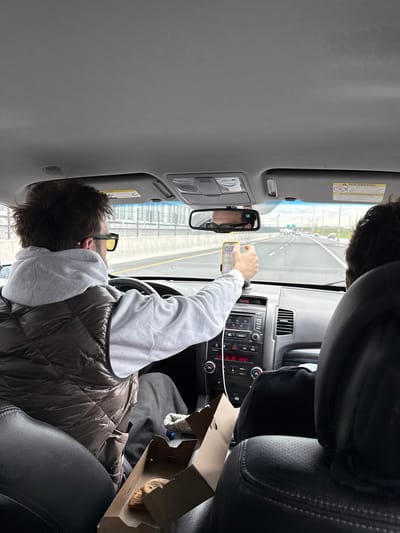 A man wearing a grey hooded sweatshirt, a brown puffer vest, and glasses is driving a car on a highway. He is looking at a phone in his hand, which is displaying a map. A cardboard box containing a baked good is partially visible on the passenger seat. The interior of the car is visible, including the dashboard, rearview mirror, and seats. Through the windshield, a highway with multiple lanes and other cars can be seen. The location appears to be a highway, but information about Brossard, Canada, is insufficient to pinpoint the exact location.