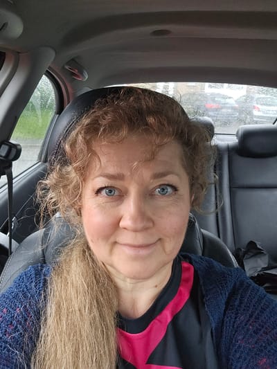 A medium close-up, eye-level selfie shows a woman inside a car, looking towards the viewer with a subtle smile. Her fair skin, bright blue eyes, and light brown/blonde curly hair are prominent. Her hair cascades over her shoulders and around her face. She wears a black top with a distinctive pink stripe and a blue, loosely knitted garment over it.
The setting is the interior of a car, with black leather seats and grey plastic trim visible around her. Through the car window behind her, visible on the left side of the frame, there are reflections and what appears to be raindrops or a wet surface, suggesting rainy or recently rainy weather outside. Blurred greenery and hints of other structures can be seen through the wet window. The overall lighting suggests it is daytime. The woman's expression is calm and pleasant. The location context provided is Philippeville, Belgium. There is no visible text in the image.