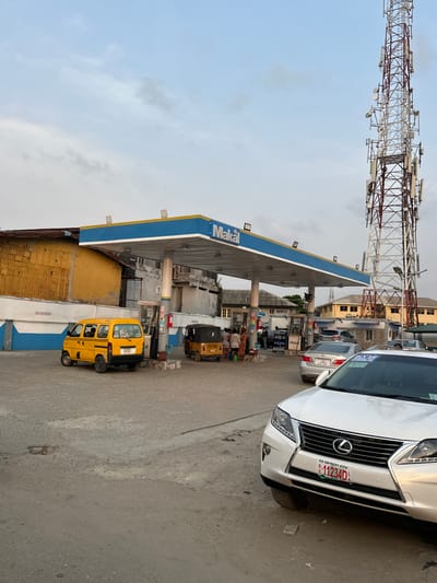 The image displays an urban scene at a "Makal" branded gas station in Lagos, Nigeria, as indicated by the license plate on a prominent vehicle. The scene is observed under an overcast sky during daylight hours.
In the foreground, a white Lexus SUV, model RX series, is partially visible on the right, facing towards the left. Its front license plate displays "LAGOS, NIGERIA" at the top, "BB SAN WALE'S AUTOS" below that, and the identifier "11234D". The ground surface is paved asphalt or concrete, showing signs of wear, including cracks and uneven patches.
The central feature is the Makal gas station, identifiable by its blue and yellow canopy supported by white pillars. Under the canopy, multiple fuel pumps are visible. To the left, a yellow minivan, possibly a Suzuki Carry or similar compact model, is parked at a pump, facing right. In the center-midground, a yellow motorized tricycle (keke napep) is also positioned near a fuel pump. Approximately 5-7 individuals, including at least one person potentially in a high-visibility uniform, are gathered around the tricycle and pumps, suggesting transactions or waiting.
Behind the Lexus SUV, a silver sedan is partially visible, appearing to queue for service. In the background, a tall, red and white lattice telecommunications tower, equipped with numerous antenna arrays, stands prominently on the right side of the frame. Surrounding the gas station are various urban structures, including a dilapidated yellow-walled building directly behind the left side of the canopy, and other residential or commercial buildings further in the background. A "NO SMOKING" sign is visible on a blue-and-white striped wall to the far left.