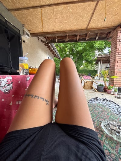 An outdoor shot features a person's bare legs and thighs, with a tattoo visible on the left thigh. The legs are positioned over a patterned blanket on a tiled floor. In the background, a table with a pink, patterned tablecloth is set with a glass jar of water and lemon, and a bowl containing food. A black bag is also on the table. The scene is set on a covered patio, with exposed wooden beams and plywood ceiling. Beyond the patio, trees and a brick structure are visible. The location is identified as Belopoptsi, Bulgaria.