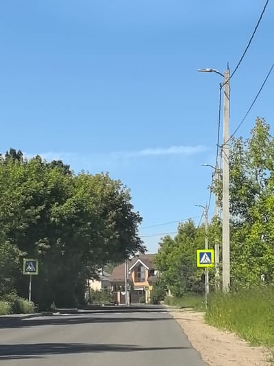 The image shows a road in the village of Afanasovo, Russia, under a clear blue sky. On the left side of the road, a pedestrian crossing sign is visible. Further down the road, houses and trees line the street. On the right side of the road, another pedestrian crossing sign is present, along with a utility pole with multiple lights and wires. The road itself is paved and appears to be in good condition. The environment is rural, with abundant greenery and a sense of quietude.