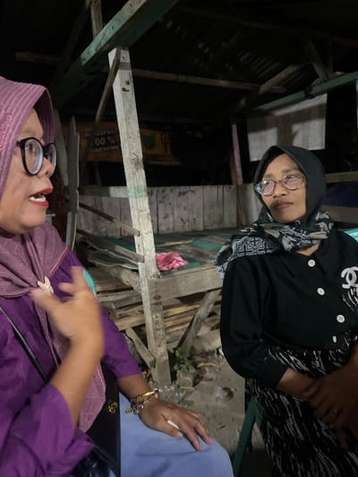 Two women are conversing at night in what appears to be an outdoor, covered structure, possibly a roadside stall or simple pavilion, in Wedarijaksa, Indonesia. The woman on the left wears a pink hijab, glasses, and a purple top adorned with a brooch. She gestures with her left hand while speaking. The woman on the right wears a black hijab and a black shirt with a white pattern and a "Chanel" logo pin. She has glasses and is looking to her left. Behind them, wooden structures and scattered debris suggest a rustic or under-construction environment. A sign with Indonesian text and a percentage is visible in the background.