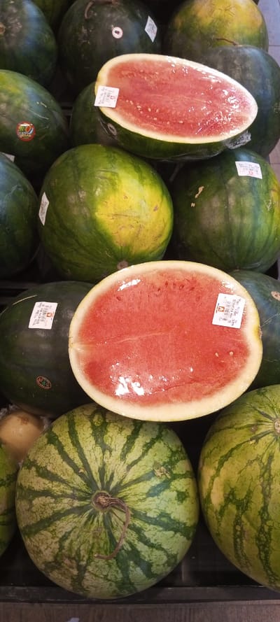 A market display of watermelons features halved fruits wrapped in plastic, showcasing their ripe, red flesh and seeds. These are surrounded by whole melons in varying shades of green, some with yellow patches and distinct stripe patterns. Price stickers are visible on several watermelons. The arrangement suggests a selection offered to customers, highlighting the quality and ripeness of the watermelons.