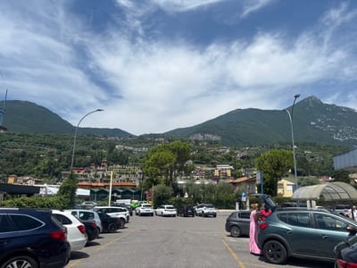 This is a wide-angle, eye-level, daytime photograph taken in a parking lot in Maderno, Italy. The scene is dominated by a row of parked cars in the foreground, with a green SUV having its rear hatch open in the middle right of the frame. A woman in a long pink dress is standing next to the open SUV hatch, facing away from the camera. A man in a red shirt is standing on the driver's side of the same SUV, with his back to the camera.
In the midground, a streetlamp with a curved arm is visible. Behind the cars and streetlamp, a hillside town with buildings and lush green vegetation stretches upwards. The town features houses with terracotta roofs and some larger, modern structures. Further in the background, majestic, tree-covered mountains rise sharply against a bright blue sky filled with wispy white clouds. The overall impression is a sunny day in a scenic, hilly location.