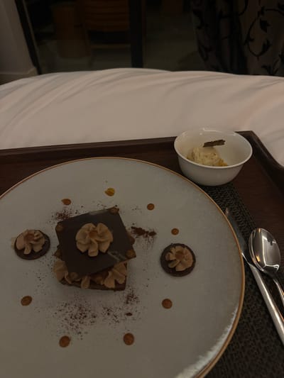 The image shows a dessert plate with a chocolate dessert on it, accompanied by a small white bowl with a scoop of ice cream. The plate is on a wooden tray, along with a silver knife and spoon. The tray is on top of a white bed sheet. The image is taken from a slightly overhead angle, and the lighting is dim. The dessert is a layered chocolate and mousse dessert, garnished with chocolate curls and a dusting of cocoa powder. The image appears to be taken from a hotel room in Ouled Jelal, Morocco. The GPS coordinates (31.664, -7.87) correspond to a location in Ouled Jelal, Morocco.