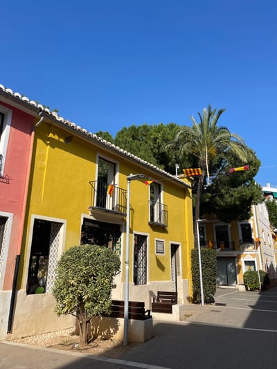The original image, taken in Picanya, Spain, is rotated 90 degrees clockwise, presenting a vibrant street scene under a clear blue sky. When viewed correctly, the scene showcases colorful architecture, lush greenery, and strong sunlight.

In the foreground, a portion of a red-orange building with white trim is visible on the left. Adjacent to it stands a prominent two-story yellow building, featuring traditional tiled roofs, windows with black metal grates or small balconies, and decorative ceramic tiles around some openings. Several red and yellow striped flags, characteristic of the Senyera (the Valencian flag), are displayed. One flag hangs from a string stretched across the street, leading towards a tall palm tree, while others are affixed to balconies. A small, square plaque with an indecipherable design or text is visible on the yellow wall.

To the right, the paved street is illuminated by bright sunlight, casting long, distinct shadows from unseen objects and the buildings themselves. Several street benches are placed along the pavement. Further down the street, another cream-colored building can be seen, also adorned with flags and featuring a large window, possibly belonging to a shop or office, though any text or logo within it is not legible.

The scene is enhanced by abundant greenery, including the large palm tree with its broad fronds, other dense green trees, and a neatly trimmed, rounded bush situated in front of one of the buildings. The sky above is a brilliant, unblemished blue, indicating a clear, sunny day. No people are visible, lending a quiet, serene atmosphere to this colorful Mediterranean street.
