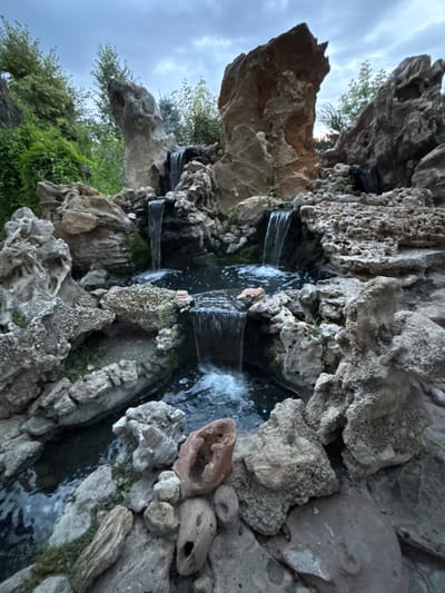 The image depicts a close-up view of a rock garden with a small waterfall. The scene is dominated by various rock formations, some porous and light-colored, others darker and smoother. Green vegetation, likely moss and small plants, is visible growing in crevices and around the base of the rocks. Water cascades down a series of rock ledges, creating small waterfalls that flow into a dark pool of water. The texture of the rocks is a prominent feature, with some appearing rough and weathered, while others have a more crystalline appearance. A few rounded stones and a reddish, irregularly shaped object are also scattered amongst the rocks. The overall impression is one of a natural, serene, and slightly weathered landscape, possibly a decorative water feature or a natural rock formation. The lighting suggests it could be daytime, possibly overcast given the soft light and cloudy sky hinted at in the background. There is no visible text in the image.