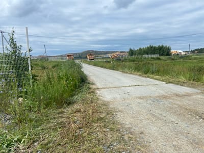 This image depicts an outdoor scene in Plast, Russia, featuring a dirt road leading into a grassy field. In the distance, several large yellow mining trucks are parked, suggesting a potential construction or mining site. The sky is overcast with gray clouds, indicating a cloudy day. Green trees are visible in the background, bordering the field. A wire fence runs along the left side of the road, with some vegetation growing through it. The overall setting appears to be rural or industrial, with an emphasis on the natural landscape and heavy machinery. There is no visible text in the image.