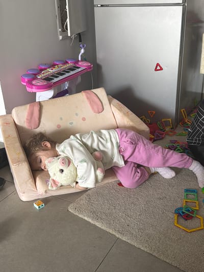 A toddler in pink pants and white socks is lying upside down in a child's chair. The child is hugging a stuffed animal pig. A toy piano is next to the chair. Scattered on the floor are magnetic toy tiles. The scene appears to be indoors, likely a home. The location is Harish, Israel.
