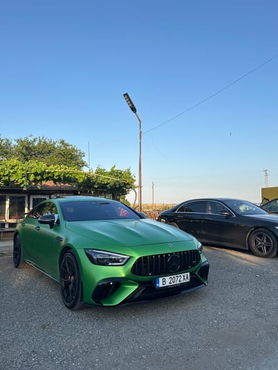 Two Mercedes-Benz vehicles are parked on a gravel lot in front of a building with a vine-covered pergola. The vehicle in the foreground is a matte green Mercedes-AMG GT 4-Door Coupe with Bulgarian license plate "B 2072 XA". It is facing the viewer at a slight angle. The vehicle in the background is a black Mercedes-Benz S-Class sedan, partially obscured by the green car. A tall metal pole with a solar-powered light fixture stands behind the green car, and overhead wires stretch across the clear blue sky. In the distance, a power line tower and some dry vegetation are visible. The location is identified as Slanchevo, Bulgaria.
