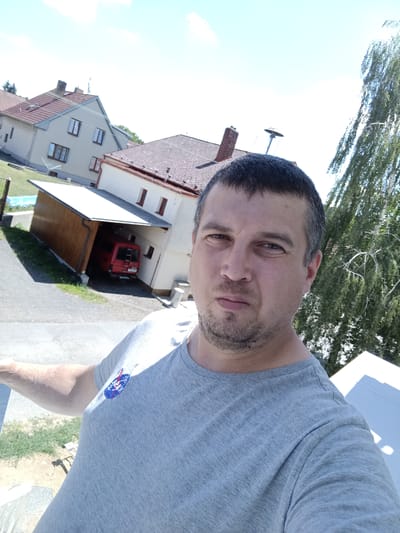 A man is taking a selfie outdoors. He is wearing a grey t-shirt with a NASA logo on the left chest. The background shows a residential area with houses and a garage. In the garage, a red van is parked. A large tree with green foliage is visible to the right of the man. The sky is bright blue with some clouds. The image appears to have been taken in Rataje, Czechia.