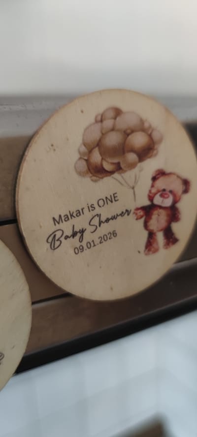 This media file shows a circular wooden ornament featuring a drawing of a teddy bear holding a bunch of balloons. Text on the ornament reads, "Makar is ONE Baby Shower" and "09.01.2026". The drawing style is whimsical and suitable for a child's celebration. The ornament is light-colored wood with a visible grain. The background is blurred, but appears to be a shelf or display surface. The lighting suggests an indoor setting, possibly a home or a shop. The overall impression is festive and celebratory, marking a child's first birthday and baby shower. The date indicates a future event. The location context is Liebenburg, Germany.