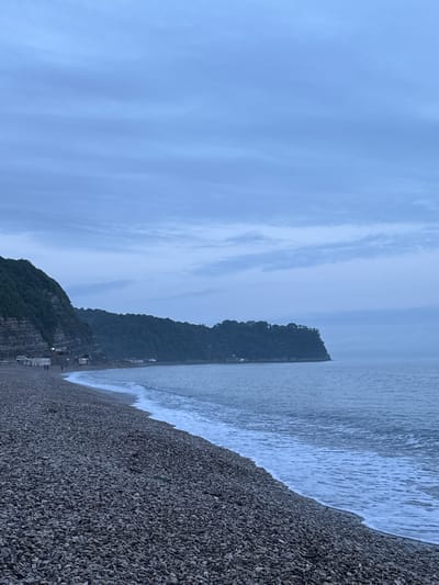 The image shows a pebble beach next to a body of water. In the background, there's a landmass, possibly a headland or a small island. The sky is overcast. It is unknown if the location is in the city of Novomikhaylovsky, Russia.