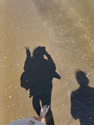 The image is a high-angle shot taken on a beach with wet sand, likely near the water's edge. Small seashells are visible scattered across the sand. Two dark shadows are cast on the sand, one resembling a person holding up a peace sign, and another, more distant shadow, is blurry, possibly belonging to another person or object. A single bare foot and the edge of a light-colored garment (possibly a dress or shirt) are visible in the lower right corner of the frame, belonging to a person taking the photo. The location, Anzio, Italy, is inferred as it is the location the photos are taken, it is the same place and time.
