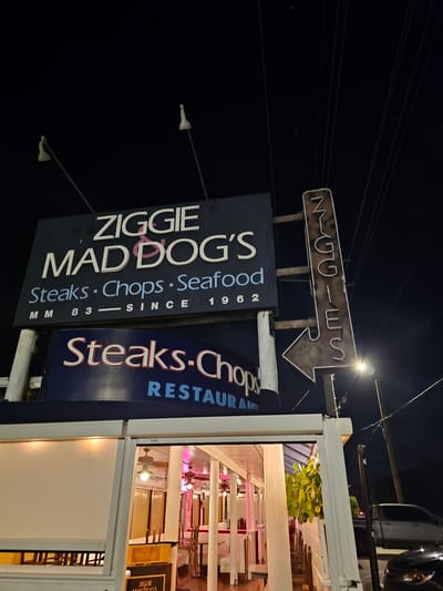 The image captures a nighttime view of Ziggie Mad Dog's, a restaurant located in Islamorada, United States of America. A large, dark blue sign with white and pink lettering reads "ZIGGIE MAD DOG'S Steaks · Chops · Seafood MM 83 · SINCE 1962". Below this, another blue sign displays "Steaks-Chops RESTAURANT" in white and red lettering. A directional arrow sign, also with neon lettering that spells "ZIGGIE", points to the left. The restaurant itself is visible through an open-air structure, with tables and chairs set up inside, illuminated by interior lighting. The scene is outdoors at night, with power lines visible against the dark sky.