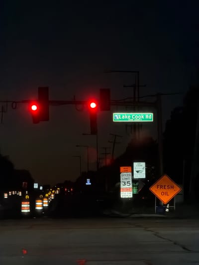 The image captures a nighttime scene at a road intersection in Buffalo Grove, United States of America. Traffic lights are illuminated red, indicating a stop. A street sign clearly reads "Lake Cook Rd" with "COOK COUNTY" below it. Several traffic signs are visible, including one indicating "WORK ZONE," "SPEED LIMIT 35," and "$250 FINE MINIMUM." Another sign warns of "FRESH OIL." Numerous orange and white traffic barrels line the road, suggesting ongoing construction or maintenance. The sky is dark, and streetlights are intermittently visible in the distance. The overall environment appears to be a suburban road setting at night, with the presence of roadwork.