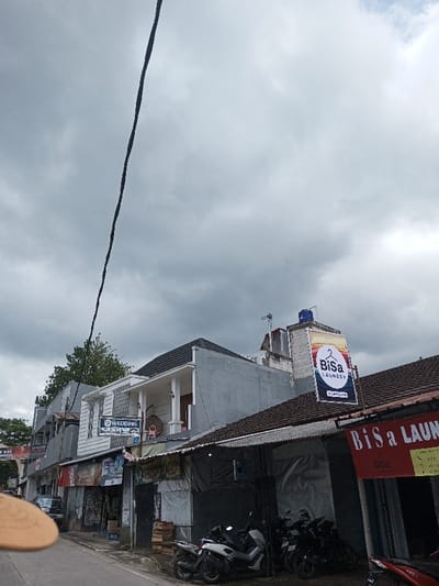 The image displays a street scene in Cibadak, Indonesia, under an overcast, cloudy sky. A dark electrical cable diagonally traverses the upper left portion of the frame. On the right side of a paved road, a row of multi-story buildings lines the street. A prominent two-story building features a grey ground floor and a white upper floor with a small, pillar-supported balcony. A rectangular sign labeled "WEDDING" is affixed below this balcony. To its right, a grey building with a dark roof displays a large vertical banner advertising "BiSa Laundry," with a blue water tank and a television antenna visible on its rooftop. Below this, a lower, single-story building with a dark corrugated roof fronts the street, displaying a partially visible red sign also reading "BiSa Laundry." Multiple motorcycles and scooters, including a white/silver model, are parked on the sidewalk area in front of these lower buildings, alongside several wooden crates. A dark minivan is partially visible on the left edge of the road. A blurry, light-colored object occupies the lower left foreground. No individuals are clearly discernible within the scene.