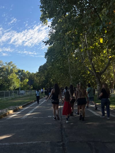 The image displays an outdoor scene on a paved pathway in San Isidro, Argentina, under a clear blue sky with scattered white clouds. Approximately 10 to 15 individuals are observed walking away from the camera on the pathway, primarily on the right side of the frame. The paved surface features double white painted lines down its center. To the left of the pathway, a grassy strip is visible, bordered by a continuous line of silver-colored temporary metal barricades. Beyond the barricades, a dense area of green trees and a tall light pole equipped with multiple floodlights are present. The right side of the pathway is densely lined with tall, mature trees exhibiting green foliage, casting partial shadows across the path. Sunlight is strong, indicating daytime. The individuals are dressed in casual attire; notable figures include a woman in black shorts with "JUICY" text and a black tank top, a child in a red dress, and another woman in a leopard-print top with black shorts. All visible individuals are in the process of walking forward along the path.