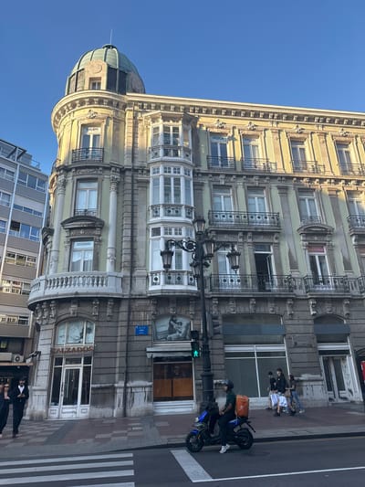 The image captures a street scene in Oviedo/uviéu, Spain, focusing on a grand, pale yellow building with an ornate facade. The building features multiple stories with balconies, arched windows, and decorative elements. A distinctive green dome caps the rounded corner of the building. On the ground floor, storefronts are visible, including one with a sign that reads "CALZADOS" (shoes).

A pedestrian crossing is in the foreground, and several people are walking on the sidewalk. One person is riding a blue electric scooter, wearing a helmet and a bright orange bag on their back, suggesting a delivery person. The sky is clear and blue, indicating fair weather. Street lamps and traffic lights are also present. The overall impression is a bustling urban environment with a blend of historic architecture and modern life.