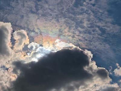 This is a photograph of a sky filled with clouds. A large, dark cloud occupies the lower right portion of the frame. Above it, and to the left, are lighter clouds that are illuminated by the sun, which is not directly visible but is implied to be behind the dark cloud. A band of iridescent colors, reminiscent of a cloud iridescence or a sun dog, is visible across the upper portion of the image, illuminating some of the smaller, higher clouds with hues of pink, yellow, and blue. The background is a deep blue sky, partially obscured by the clouds. The image was taken in Wilnsdorf, Germany.