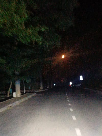 The image depicts an asphalt road at night, illuminated by a crescent moon visible in the dark sky. Trees with dark foliage line the left side of the road, casting shadows. White dashed lines mark the center of the road. Several streetlights and possibly illuminated signs are visible in the distance, creating points of light against the darkness. The environment suggests a street or road in a semi-urban or suburban area.