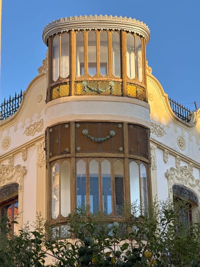 The image captures an ornately decorated building facade in Vinaròs, Spain, featuring a curved, enclosed balcony. The balcony has a series of large windows with wooden frames, some with decorative metal grilles featuring floral designs. The building's exterior is adorned with intricate plasterwork and moldings. Trees are partially visible on the right side of the image. The sky is clear and blue.