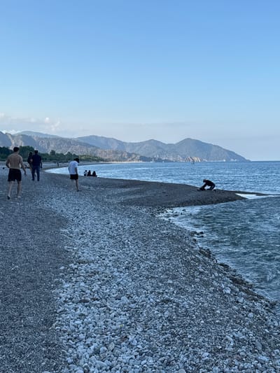 This image captures a serene coastal scene in Kumluca, Turkey, featuring a wide pebble beach, a calm sea, and distant mountains under a clear, bright blue sky. The lighting suggests it is late afternoon or early evening.
Several individuals are present on the beach. In the foreground, three men walk along the smooth, grey pebble shore, heading away from the viewer. The closest man is shirtless, wearing dark shorts, while the other two wear t-shirts and shorts. Further down the beach, a small group of people can be seen near the waterline.
To the left, closer to the water, a lone figure dressed in dark clothing, possibly a wetsuit, is seen bending over at the edge of the sea, seemingly engaging with the shallow water or a small object. The sea is calm, displaying a deep blue hue with gentle ripples.
In the background, a majestic range of mountains stretches across the horizon, their peaks softened by a light haze, characteristic of a coastal landscape. The sky above is vast and mostly cloudless, contributing to the tranquil atmosphere. No visible text is discernible in the image.