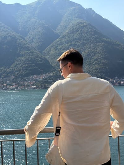 A man stands facing away from the camera, leaning on a railing, looking out at a body of water and a mountain range dotted with buildings. The man is wearing a light-colored, long-sleeved collared shirt and sunglasses. A strap from a bag is visible across his back and shoulder. The water is a bright blue with sunlight reflecting off its surface. The mountains in the background are covered in green vegetation, with a town or village nestled along the shoreline. The sky is clear and blue. The scene appears to be a scenic overlook in Pognana Lario, Italy.