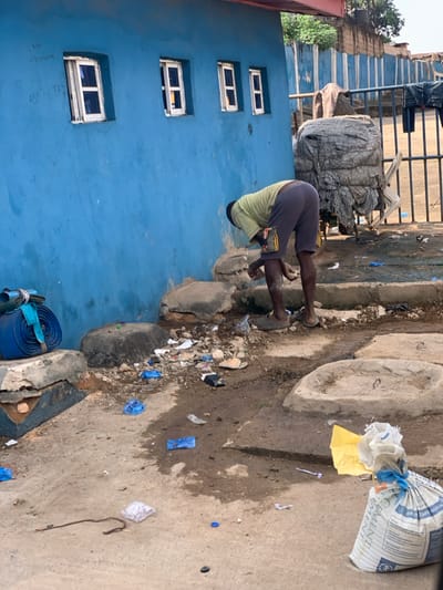 The image depicts an outdoor scene in Ikpoba, Nigeria. An adult male, wearing a light green short-sleeved shirt, dark grey shorts, and light-colored flip-flops, is positioned in the center-right, bent forward at the waist. His hands are near a small, light-colored protrusion at the base of a vibrant blue-painted building wall, suggesting an interaction with a water source or fixture. An object, possibly a small bag or phone, is visible attached to his right thigh.

The blue building wall features four rectangular, white-framed windows evenly spaced across its upper section. Faint writing and numbers, including "Felix" and "081..." sequences, are visible on the upper left portion of the wall.

The ground in the foreground and midground is a mix of dirt and concrete, heavily soiled and littered with various refuse. Visible debris includes multiple blue plastic wrappers, a yellow plastic sheet, a black bottle cap, small rocks, and general trash. Patches of dampness are present on the ground. Several irregular concrete blocks or raised platforms are also visible on the ground.

To the far left, a large blue hose, tightly rolled and secured, rests on a concrete block. In the right foreground, a white woven bag, clearly marked "CEMENT" and "MADE IN NIGERIA," tied with a blue string, stands upright.

In the background, beyond the person and the blue wall, a metal fence with vertical bars is visible. Behind the fence, there is a large, irregularly shaped object covered in grey tarp or fabric, possibly a bundled load on a cart or structure. Further back, a reddish-brown wall and some distant vegetation are discernible. The scene is illuminated by natural daylight.