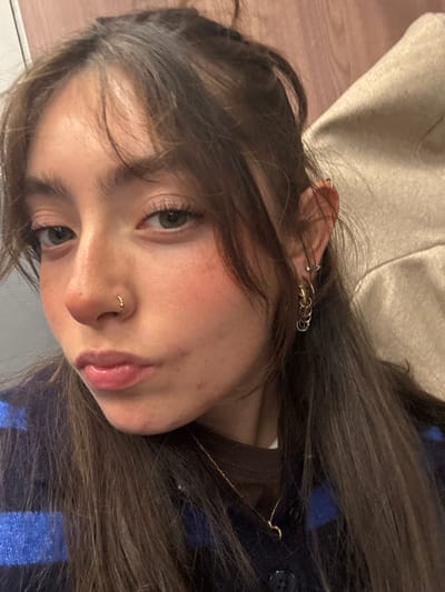 A young woman poses, looking into the camera. She has long brown hair with bangs, styled casually. Her complexion is fair, and she has a nose ring and multiple earrings. She wears a brown top and a blue and black plaid jacket. The background is simple, with a glimpse of a beige fabric in the top left corner.