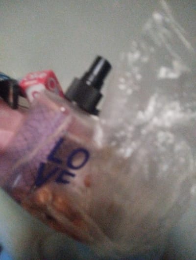 The scene is located in Kaninkon, Nigeria. The image displays a blurry, out-of-focus view of several items on a plain, light-colored surface. A translucent, rectangular bottle, approximately centered, features a black spray nozzle and partial dark blue lettering "LOVE" on its side. The bottle contains a reddish-brown liquid with visible particulate matter. To the immediate right and slightly behind this bottle, a clear plastic bag holds a white, granular or crystalline substance. A small, partially visible red and white object is situated behind the bottle on the left. No individuals, actions, or interactions are visible.
