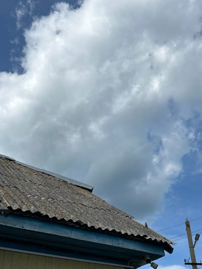 This low-angle, outdoor shot captures a cloudy sky over a corrugated roof. The sky is a mix of deep blue and lighter grey clouds, with some areas appearing bright white, suggesting the sun is present but diffused. In the distance, a small, dark bird is visible in flight against the clouds.
The foreground is dominated by the edge of a weathered, grey corrugated roof, which appears to be part of a building. A blue metal gutter runs along the edge of the roof. To the right of the roof, a portion of a pale yellow building is visible. Above and to the right, a wooden utility pole with two lights and electrical wires cuts across the upper portion of the frame.
The scene conveys a sense of an overcast day, possibly in the afternoon, given the lighting. There are no people visible. The setting appears to be outdoors, likely in a rural or semi-rural area given the presence of the utility pole and the type of roofing. The overall mood is neutral, focusing on the architectural elements and the sky. No text is visible in the image.