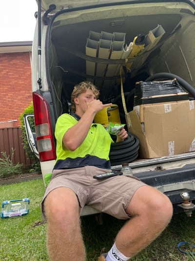 A young man in a neon yellow work shirt and tan shorts reclines in the back of a white van. He points his finger up while holding a bag of chips. The van's cargo area is filled with equipment, including pipes and boxes. The van's open rear doors and a glimpse of a brick wall and green lawn are visible outside the vehicle. A clear toolbox sits on the grass beside the van.